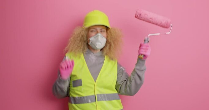 Joyful female construction worker dances happily with paint roller jumps and moves actively wears protective helmet uniform and safety clothes poses against pink background ready for repair.