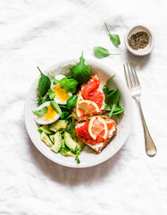 Bowl of green salad, avocado, boiled egg and sandwiches with rye bread, cream cheese and salmon on a light background, top view. Delicious and healthy breakfast, brunch