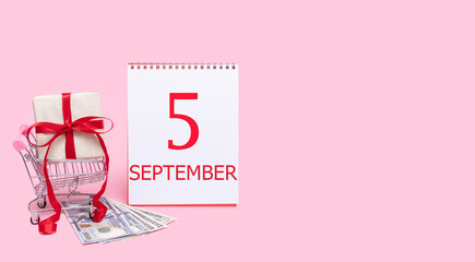 A gift box in a shopping trolley, dollars and a calendar with the date of 5 september on a pink background.