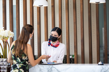 beautiful young asian woman receptionists working at a reception desk and holding key card or passport to customer. People wearing protective face mask prevent covid-19 virus - Powered by Adobe