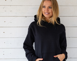 A smiling blonde is standing in a black sweatshirt on a background of white boards, in front of camera. There is a blank space on the garment for a design, logo or inscription. Mockup for clothes. - 440534843