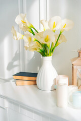 Soft home decor, white jug, vase with white and yellow beautiful flowers on a white wall background and on a wooden shelf. Interior.