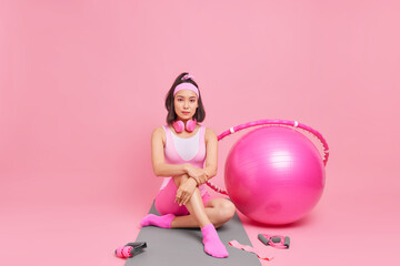 Serious brunette fit Asian sportswoman in sportsclothes sits on fitness mat after workout surrounded by different sport equipment isolated over pink background. Sport and motivation concept.