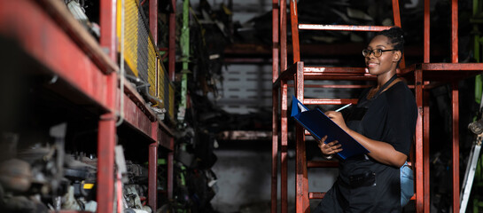 Plus size black female worker inspecting old car parts stock while working in automobile large warehouse