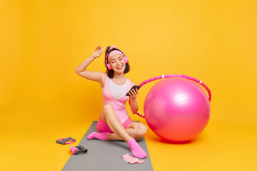Indoor shot of carefree young Asian woman enjoys favorite playlis holds mobile phone wears stereo headphones dressed in activewear sits on mat trains at home uses fitness ball hula hoop expander