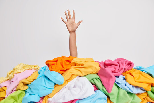 Unrecognizable human raises arm reaches out heap of colorful unfolded clothes busy doing wardrobe cleaning isolated over white background. Woman buried under cluttered clothing items. Decluttering