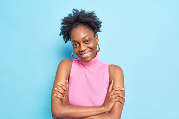 Portrait of good looking woman with dark skin curly hair smiles pleasantly keeps arms folded tilts head dressed in pink shirt isolated over blue background. Human emotions and sincere feelings concept