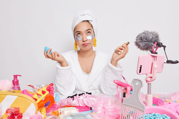 Serious thoughtful Asian lady holds bottle of foundation and sponge undergoes beauty treatment applies patches under eyes recods video for her blog uses phone camera reviews cosmetics at home