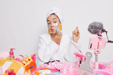 Serious woman looks attentively at camera of mobile phone applies foundation on face with sponge beauty patches under eyes wears soft bathrobe towel does video recording recommends cosmetic products