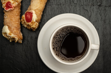 Top view and close-up cup of coffee accompanied with two cannoli on black stone slab