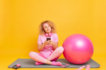 Pretty dreamy curly female model in good physical shape poses crossed legs on fitness mat takes...