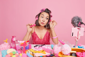 Asian female blogger applies mascara reviews make up cosmetic products films process does online video translation from home wears hair rollers dressed in fashionable pink dress poses indoor