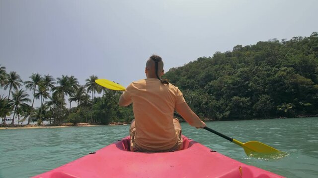 Young man with sunglasses and hat rows pink plastic canoe along sea against green hilly islands with wild jungles. Traveling to tropical countries. Strong guy is sailing on kayak in ocean, back view.