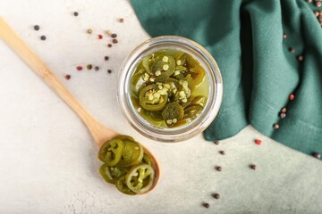 Glass jar and wooden spoon with canned jalapeno on light background