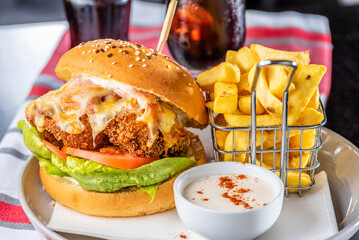 A chicken parma burger with lettuce and tomato on a white plate with french fries in a wire basket...