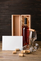 Box with bottle of wine, glasses and gift card on wooden background