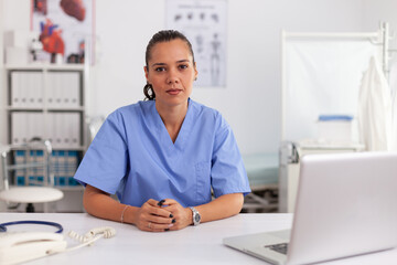 Portrait of pretty medical nurse smiling at camera in hospital office wearing blue uniform. Healthcare practitioner sitting at desk using computer in modern clinic looking at monitor, medicine.