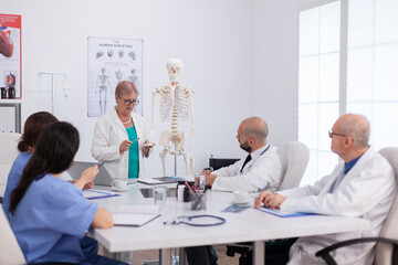 Obraz premium Physician senior woman holding bone structure discussing human structure using body skeleton presenting healthcare treatment. Hospital group working at medical expertise in meeting room