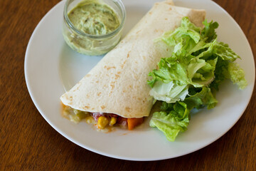 A burrito filled with chili sauce on a plate with some green lettuce and a small bowl of guacamole on the side