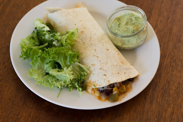 A burrito filled with chili sauce on a plate with some green lettuce and a small bowl of guacamole on the side