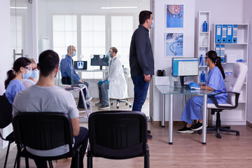 Patient visiting hospital clinic during covid 19 pandemic, assistant wearing visor checking appointment in reception area. People waiting for consultation sitting in hallway respecting social distance
