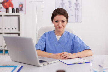 Portrait of medical practitioner nurse looking into camera smiling sitting at desk in conference meeting room. Confident physician asisstant analyzing pills treatment expertise on laptop computer