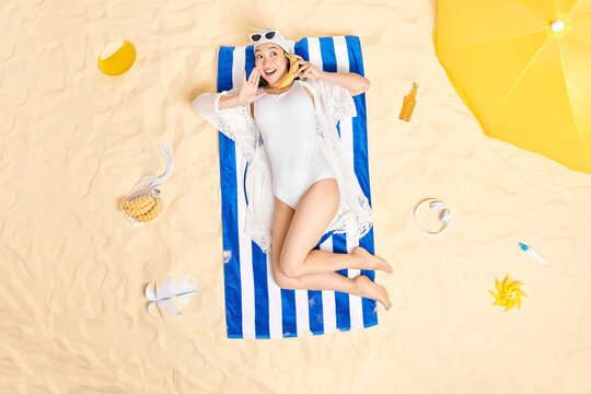 Pleased young Asian woman in white bikini and sunhat keeps banana near ear pretends calling someone has happy mood lies on towel poses at beach enjoys relaxation time during summer holidays.
