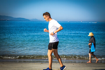 young man running along the beach in pandemic time with woman in the background wearing medical mask as prevention