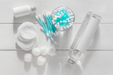 Glass with cotton swabs, pads, balls and bottles of cosmetic products on light wooden background