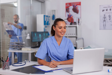 Smiling nurse using laptop computer and writing notes on clipboard in hospital office with doctor in the background. Health care physician using computer in modern clinic looking at monitor,