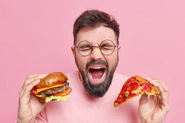 Emotional bearded man eats junk food shouts loudly keeps mouth widely opened holds burger and slice...
