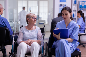 Assistant taking notes on clipboard about patient madical problems waiting for specialist doctor sitting on wheelchair in waiting room of hospital clinic. Nurse explaining medical procedure to woman