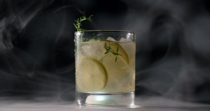 Cold glass with cocktail is spinning slowly in the smoke on the dark background, alcohol drinks and beverage, non-alcohol cocktail, object video of lemonade, 4k Prores HQ
