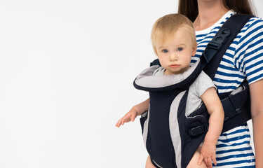 Unrecognizable woman holding caucasian adorable blond child looking at camera in soft structured baby carrier backpack indoors on white gray background.Kid and parent care concept.Copy space for text