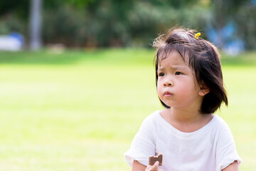 Asian child eating chocolate wafers on natural green laws background. Cute girl holding sweetmeat on her hand. Baby age 4 years old.