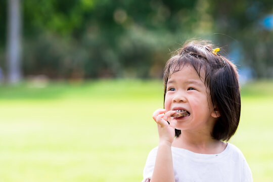Asian child eating chocolate. Cute girl biting delicious wafers. On green laws nature background. Summer or spring time. Happy kid aged 4 years old.