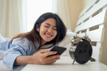 Young Asian woman smiling after waking up and holding smartphone on bed at morning