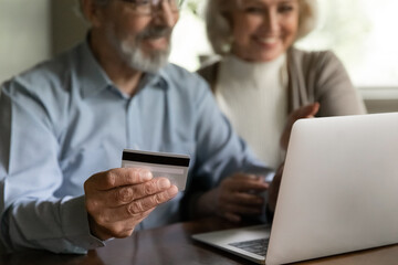 Crop close up of happy mature man and woman buyer shopping online on laptop use credit debit card. Old couple spouses clients pay buy on internet on computer from home. Secure banking concept.