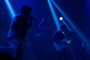 Silhouette of young Asian pop rock singer and band performing with full spectrum of light and sound at the concert. Blue light background