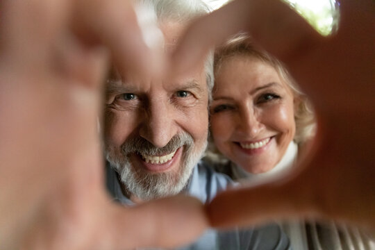 Crop Close Up Portrait Of Happy Mature Man Woman Make Heart Love Hand Gesture Sign Pose For Selfie Together. Smiling Old Couple Spouses Have Fun Take Self-portrait Picture Show Family Care Bonding.