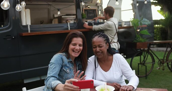 Multiracial women having fun taking photos with mobile phones at food truck restaurant outdoor - Summer and friendship concept