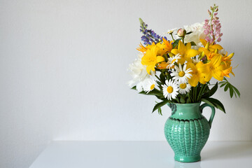 Summer bouquet in a jug of garden and wild flowers on a white table against a white wall. Arrangement consists of a bright yellow lilies, daisies, lupines. Selective focus, copy space.