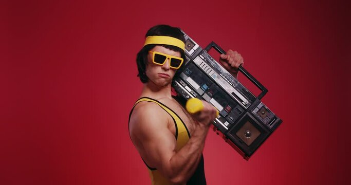 Funny retro athlete man in yellow tracksuit and sunglasses from 80's with ghettoblaster makes the workout isolated on red background. Retro sport hipster show off and posing in front of camera.