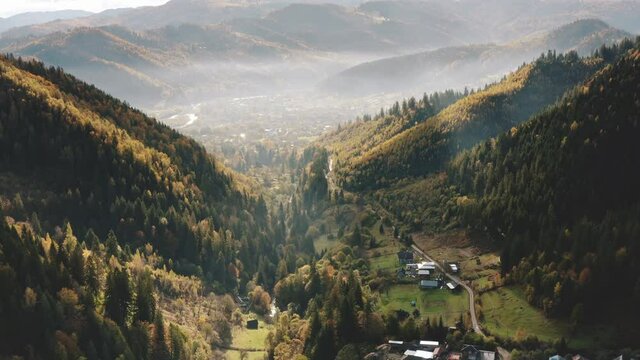 Mountain village in canyon aerial view. Green forest hills, rural road, gog and sun beams. Nature background. Travel destinations. Beautiful wild landscape. Drone high flight over Carpathians, Europe