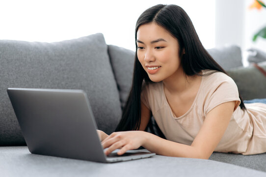 A cute positive asian girl lies at home on the couch on her stomach, uses a laptop, browses the Internet, chatting on social networks, watches an online webinar, writing on email, smiling
