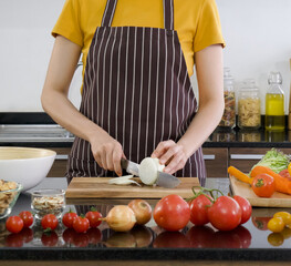 Young chef slices onion into pieces on a wooden chopping board. The kitchen counter full of various kinds of vegetables.