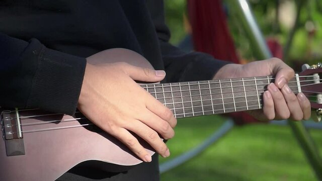 The young guitarist plays a classic wooden ukulele. A musician plays an acoustic ukulele in nature in the morning. Close-up shot.