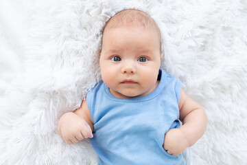 portrait of a cute baby boy three months old in a blue bodysuit on a white bed at home
