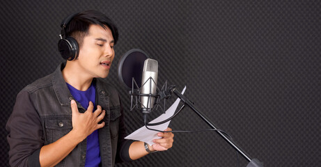 Young asian man with headphones sing while holding lyrics in his hand in front of black...