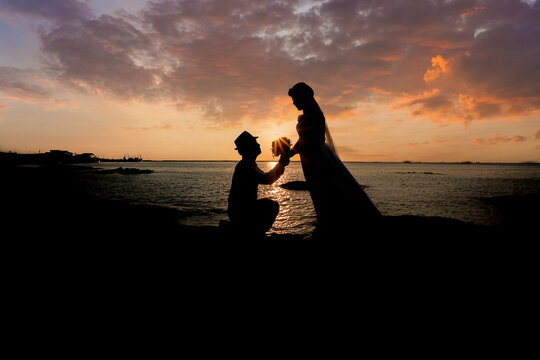 A silhouette of a young man, down on one knee and holding a bouquet, proposing to his girlfriend. will you marry me images. Young couple in love at beautiful sunset.